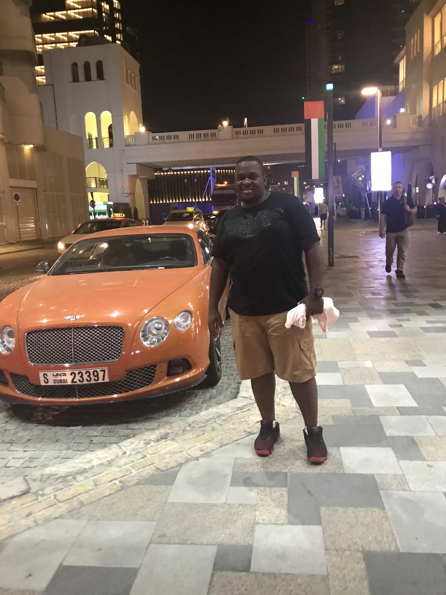 Asong beside an orange Bentley on a lit Dubai street at night