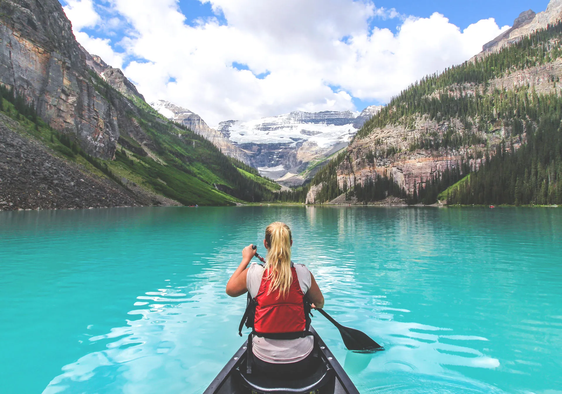 Traveller paddling a red kayak across a turquoise mountain lake under snow-capped peaks